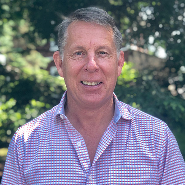Dan Cressman wearing a checkered blue, white, and red button down shirt standing in front of trees at D.E. Cressman Insurance Agency Inc. in Allentown, PA.